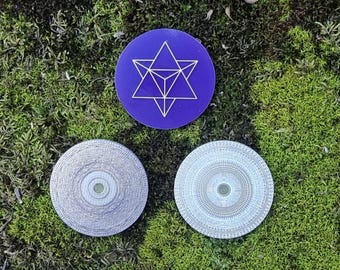 May include: Three circular objects on a bed of green moss. One is a deep purple with a white geometric star design. The other two are silver with intricate circular patterns. The objects are likely coasters or decorative pieces.