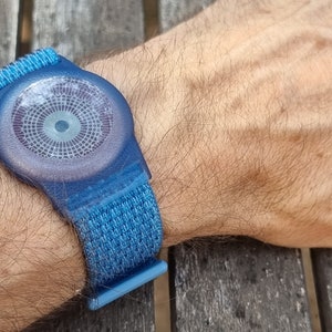 May include: A blue wristband with a circular, textured, grey face. The face has a small, white circle in the centre.