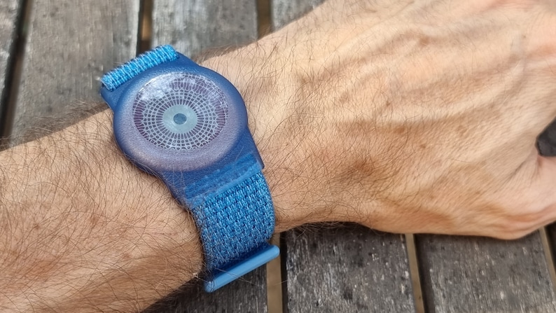 May include: A blue wristband with a circular, textured, grey face. The face has a small, white circle in the centre.