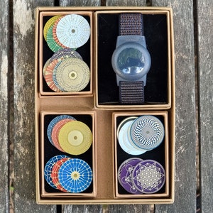 Peut inclure: Une boîte rectangulaire en carton contenant quatre compartiments. L'un contient un appareil circulaire gris foncé avec un bracelet en tissu marron. Les trois autres compartiments contiennent des piles de disques circulaires aux motifs géométriques colorés.