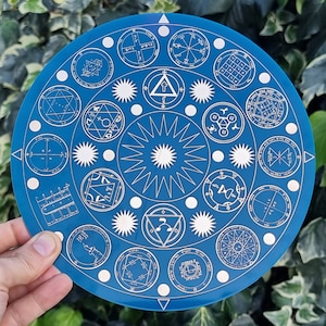 Op de afbeelding: Een blauwe ronde schijf met witte symbolen en ontwerpen. De schijf heeft een centraal stervormig patroon en is omgeven door een ring van kleinere symbolen.