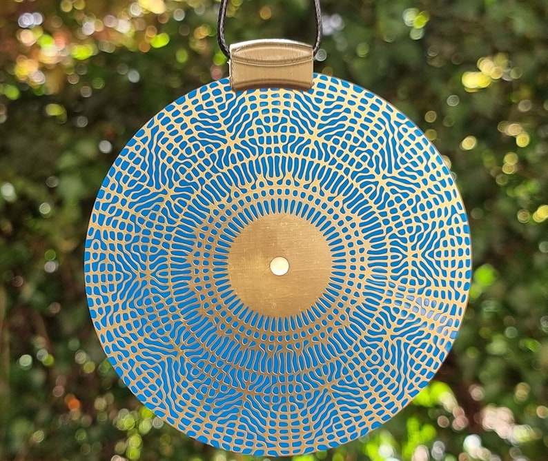 May include: A round, blue and gold pendant with a geometric pattern. The pendant is made of metal and has a cord loop at the top.
