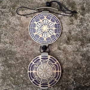May include: Two round, decorative metal pendants with a geometric pattern. The pendants are gold with a purple design and are connected by a black cord.