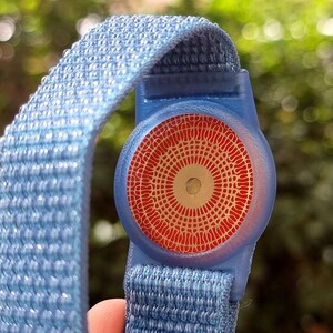 May include: A blue wristband with a red and silver circular tag. The tag has a geometric pattern.
