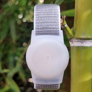 May include: A white, round, wearable device with a gray fabric band. The device has a smooth, slightly textured surface and a small, circular indentation in the center.