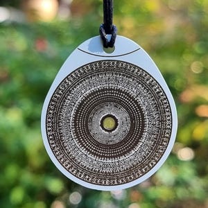 May include: A pendant necklace with a circular design. The pendant is a light color with a black cord. The pendant has a detailed, concentric circular pattern in brown and white. The background is blurred green.
