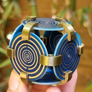 Peut inclure: Un cadre en métal doré maintient ensemble six panneaux circulaires bleus et dorés avec un motif de cercles concentriques. Les panneaux sont disposés en forme de cube.