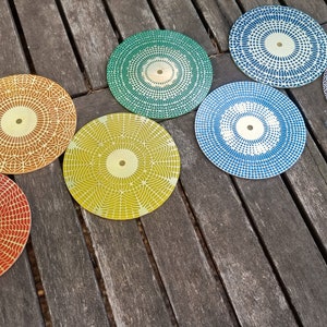 Puede incluir: Siete posavasos redondos con un patr&oacute;n geom&eacute;trico en tonos de rojo, naranja, amarillo, verde, azul y gris. Los posavasos tienen un centro dorado.