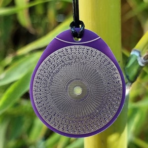 May include: A purple pendant with a silver, circular design featuring a mandala-like pattern. The pendant is on a black cord.