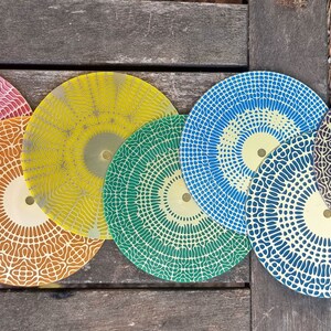 Puede incluir: Seis platos decorativos redondos con intrincados patrones geom&eacute;tricos en varios colores, incluyendo rojo, amarillo, verde, azul y negro. Los platos tienen un centro dorado y est&aacute;n dispuestos en una fila sobre una superficie de madera.