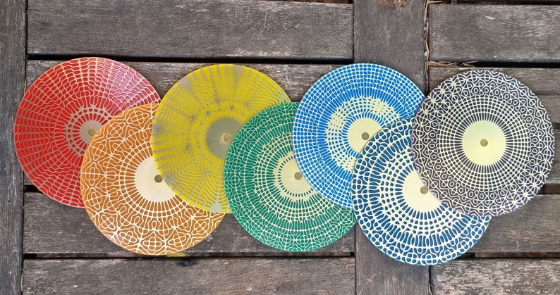 Puede incluir: Seis platos decorativos redondos con intrincados patrones geom&eacute;tricos en varios colores, incluyendo rojo, amarillo, verde, azul y negro. Los platos tienen un centro dorado y est&aacute;n dispuestos en una fila sobre una superficie de madera.