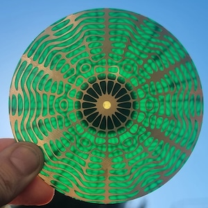 Puede incluir: Un disco decorativo redondo, verde y dorado con un patrón concéntrico de círculos y ondas. El centro del disco tiene un diseño de estrella dorada.