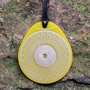 May include: A yellow pendant with a circular design and a smaller circle in the center. The pendant is hanging from a black cord.