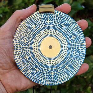 May include: A round, gold-colored pendant with a blue and gold geometric pattern. The pendant is hanging from a black cord.