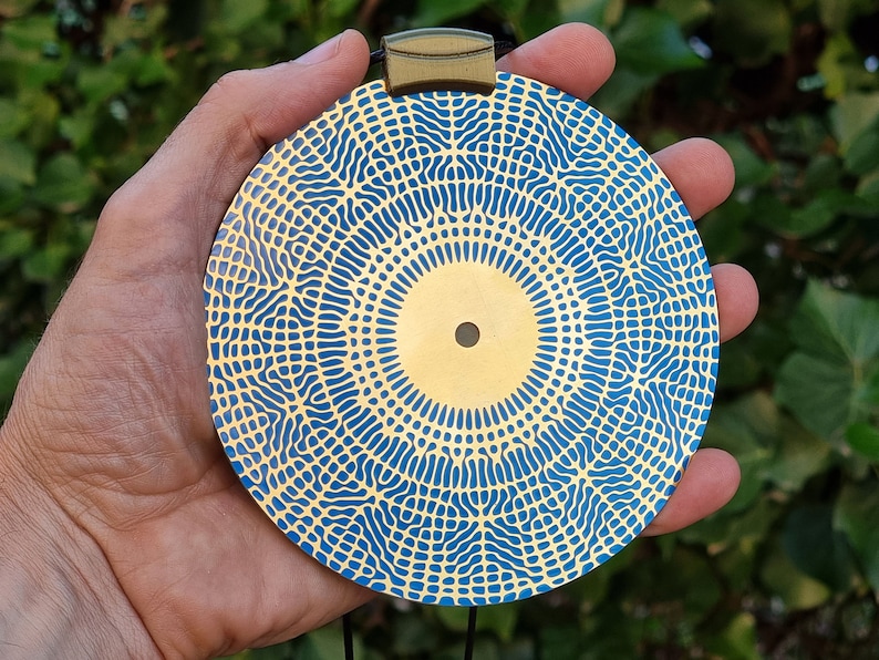May include: A round, gold-colored pendant with a blue and gold geometric pattern. The pendant is hanging from a black cord.