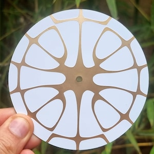 May include: White circular disc with a gold geometric pattern. The pattern resembles a stylized flower or starburst.