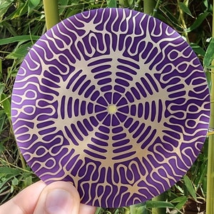 May include: A circular mandala design in purple and gold with intricate patterns. The mandala is made of a smooth, reflective material.