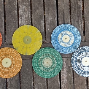 Puede incluir: Siete posavasos redondos decorativos con diferentes patrones geom&eacute;tricos en rojo, amarillo, azul, verde y negro. Los posavasos tienen un centro dorado.