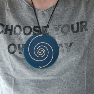 May include: A blue circular pendant with a white spiral design hangs from a black cord. The pendant is worn by a person wearing a grey t-shirt with the text "CHOOSE YOUR OWN WAY" printed on it.