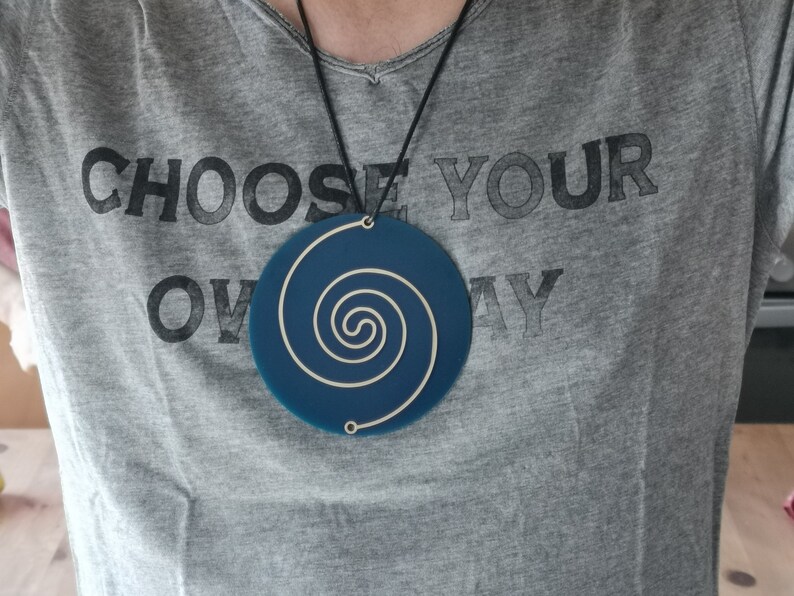 May include: A blue circular pendant with a white spiral design hangs from a black cord. The pendant is worn by a person wearing a grey t-shirt with the text "CHOOSE YOUR OWN WAY" printed on it.
