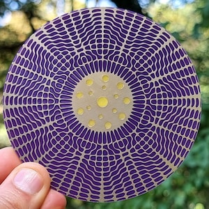 May include: A round coaster with a purple and gold geometric design. The coaster features a central circle with yellow dots and a radiating pattern of curved lines. The design is held by a hand against a blurred green background.