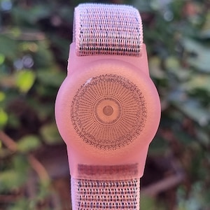 May include: A pink smartwatch with a circular face and a metallic mesh strap. The watch face has a detailed, patterned design. The strap is silver in colour.