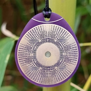 Può includere: Un ciondolo viola con un designo circolare argentato con un motivo geometrico. Il ciondolo è su un cordoncino nero.