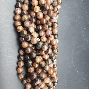 May include: Strand of round beads in shades of brown, tan, and black. The beads have a polished, glossy finish and vary in color and pattern. The beads are strung together, creating a necklace or bracelet.