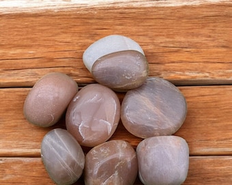 May include: A group of seven smooth, light brown and white stones with a slightly iridescent sheen. The stones are arranged in a circle on a wooden surface.