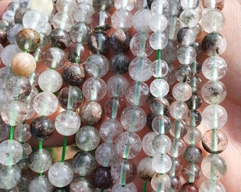 May include: A strand of clear quartz beads with green and brown inclusions. The beads are round and have a smooth, polished finish. The strand is strung on a green thread.