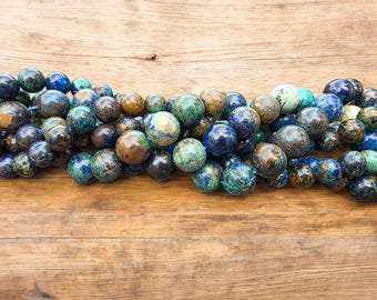 May include: A collection of round, polished beads in shades of blue, green, brown, and gold. The beads are strung together, creating a textured and colorful display. The beads are resting on a wooden surface.