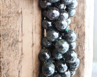 May include: A strand of dark, glossy, round beads, possibly gemstones, strung on green thread. The beads have a reflective surface, with hints of green and blue. The beads are displayed against a weathered, light brown wooden background.