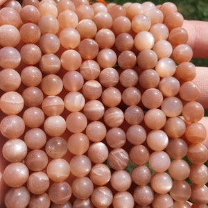 May include: A collection of peach-toned sunstone beads. The spherical beads have a polished, reflective surface. They are strung on a thin, orange cord. The beads have a warm, earthy hue, with a subtle shimmer, ideal for jewellery making.