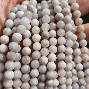 Peut inclure: Un brin de perles d'agate blanche avec une finition lisse et polie. Les perles sont rondes et légèrement translucides, révélant de subtils motifs gris et blancs à l'intérieur de la pierre.