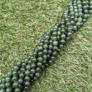 Puede incluir: Hebras de cuentas de jade verde oscuro, pulidas y redondas. Las cuentas están ensartadas, creando una forma fluida y orgánica. Las cuentas son de un color verde oscuro, con un acabado liso y brillante. Las cuentas se muestran sobre hierba verde.