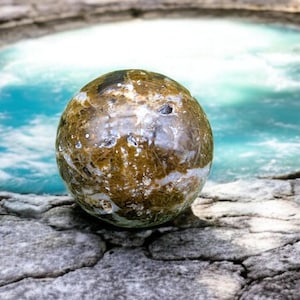 Peut inclure: Une sphère d'agate ronde, brune et blanche avec une surface lisse et polie. La sphère est posée sur une surface texturée, grise.