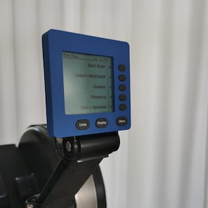 Pode incluir: Um monitor de desempenho digital azul para máquina de remo com uma tela quadrada exibindo um menu com opções incluindo Just Row, Select Workout, Games, Memory e More Options, com cinco botões laterais e três botões inferiores.