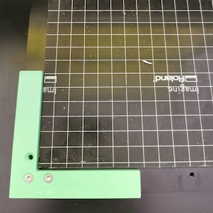 以下が含まれることがあります： 2つのネジ穴のある緑色のプラスチック製のL字型の定規。定規は、グリッドパターンが描かれた黒いカッティングマットの上に置かれています。マットには、「Imagine」と「Roland」の文字が印刷されています。