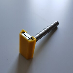 May include: A yellow plastic safety razor with a silver metal handle. The razor is designed to be used with disposable razor blades.