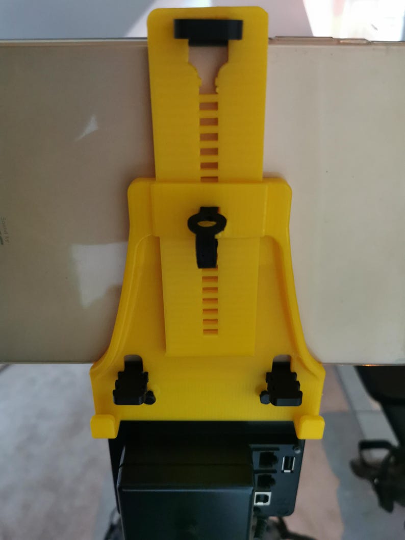May include: A yellow 3D printed phone holder with adjustable height and a black phone clip. The holder is attached to a black device with multiple ports.
