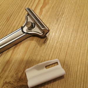 May include: A silver scraper tool with a textured blade and a white plastic handle. The tool is designed for removing wallpaper or other materials.