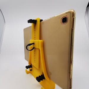 May include: A yellow 3D printed tablet stand with a black adjustable clamp. The stand is designed to hold a tablet in a vertical position.