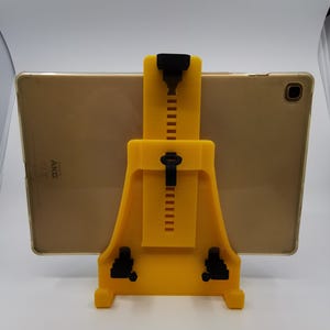 May include: A yellow plastic tablet stand with black adjustable arms. The stand is holding a gold tablet with the text "AKG" on the back.