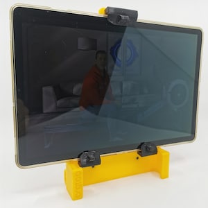 May include: A yellow 3D printed tablet stand with black clips to hold a tablet in place. The tablet is displaying a video on a screen.