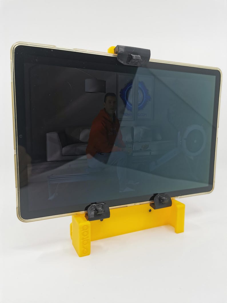 May include: A yellow 3D printed tablet stand with black clips to hold a tablet in place. The tablet is displaying a video on a screen.