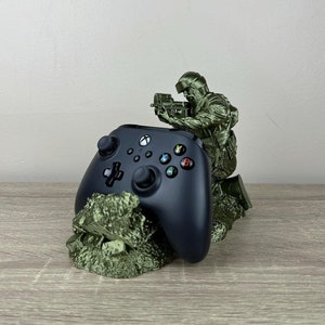 May include: A green plastic figurine of a soldier holding a gun, standing on a green rock base. A black Xbox controller is resting on the base.