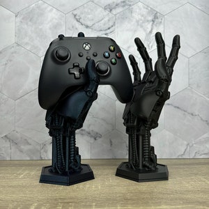 May include: A black video game controller is held by a 3D printed robotic hand. The robotic hand is on a black base. There is a second robotic hand on a black base to the right of the first.