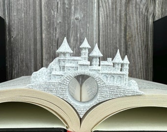 May include: A white 3D printed castle bookmark shaped like a bridge, with a book open underneath. The bookmark is made to look like stone with a rough texture.