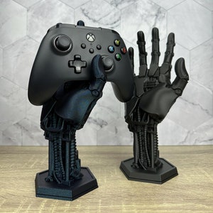 May include: A black 3D printed robotic hand holding a black video game controller. The hand is on a hexagonal base. There is a second robotic hand on a hexagonal base to the right of the first hand.