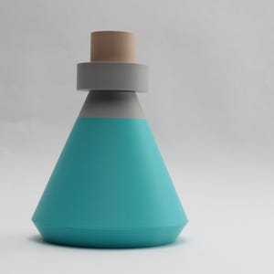May include: A teal-colored, geometric bottle with a gray band and a tan stopper. The bottle has a conical shape and a flat base. The design is minimalist and modern, suitable for home decor or as a unique container.